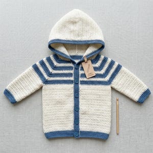 Può includere: Una giacca con cappuccio all'uncinetto color crema e blu con chiusura a bottoni. La giacca presenta strisce e finiture blu e un'etichetta con la scritta "JAMES HOOK". Un uncinetto è posto accanto alla giacca.