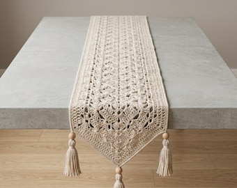 May include: Cream-colored, crocheted table runner with a detailed openwork design. The runner has a rectangular shape and is adorned with tassels and wooden beads at each end. The runner is displayed on a gray table.
