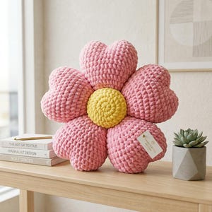Könnte beinhalten: Ein blumenförmiges Dekokissen aus grobem, weichem rosa Garn mit einem leuchtend gelben, kreisförmigen Mittelteil. Das Kissen besteht aus fünf herzförmigen Blütenblättern, die um den Kern angeordnet sind. Ein kleines Etikett hängt an einem Blütenblatt.