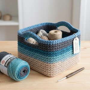 May include: A handmade square basket in shades of blue, gray, and beige, filled with yarn and small decorative stones. A ball of blue and gray macrame cotton yarn and a crochet hook are next to the basket. The basket has handles and a tag that says "Handmade Square Basket."