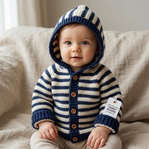 May include: A baby wearing a navy blue and cream striped hooded sweater with wooden buttons. The sweater has matching navy blue cuffs and a tag that reads "JAMES HOODIE" and "6-12 Months". The baby is sitting on a neutral-colored surface.