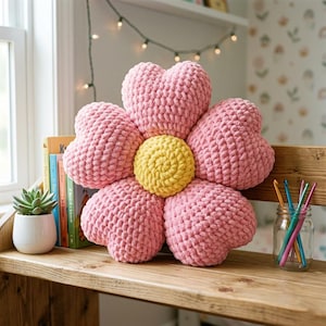 May include: A large, pink, flower-shaped pillow with a yellow center. The pillow is made of a chunky, textured yarn and sits on a wooden shelf. A small succulent in a white pot and books are to the left.