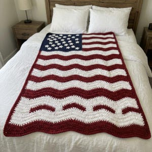 May include: A handmade crochet blanket in the style of the American flag. The blanket features a blue square with white stars in the upper left corner, with red and white stripes. The blanket is displayed on a white bed.