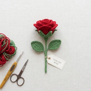 Op de afbeelding: Een handgemaakte gehaakte roos in felrood, met groene bladeren en stengel. Een label leest "CROCHET ROSE". De knutselbenodigdheden omvatten rood en groen garen, een haaknaald en een kleine schaar. De roos is een decoratief item.