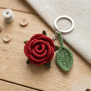 May include: A crochet keyring featuring a deep red rose flower with dark green leaves attached to a silver metal split ring. The rose is crafted with layered petals and is accompanied by a single, textured, pointed green leaf charm.
