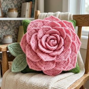 May include: A square throw pillow featuring a large, three-dimensional crocheted pink flower with layered petals centered on a green base. The design includes two crocheted green leaves extending from the side of the flower, all crafted with yarn.