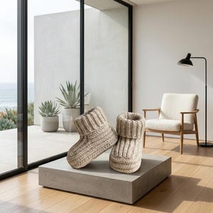 May include: A pair of beige knitted slippers with a ribbed cuff, displayed on a gray concrete block. The slippers are made of a thick, textured yarn. The background includes a modern chair, a floor lamp, and a view of the ocean.