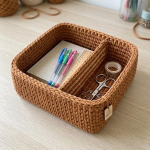 May include: A brown, crocheted desk organizer basket with a rectangular shape and internal dividers. The basket contains pens, a ruler, scissors, tape, and a notebook. A small tag reads "Desk Basket".