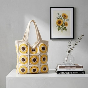 May include: A cream-coloured crochet tote bag with a sunflower pattern. The bag features yellow and brown sunflower motifs on the front and sides. The handles are also cream-coloured. A framed sunflower print and books are in the background.