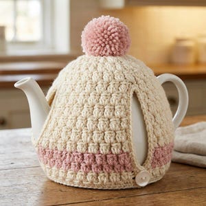 May include: A white ceramic teapot covered by a cream-colored crochet tea cozy. The cozy features a textured stitch pattern, a horizontal dusty pink stripe near the base, a dusty pink pom-pom on top, and a single white button closure at the side.