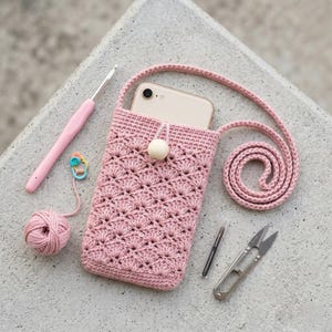 May include: A handmade pink crochet phone case with a matching strap, holding a smartphone. A crochet hook, yarn ball, stitch marker, small scissors, and a needle are arranged around the case on a concrete surface. The case has a wooden bead closure.