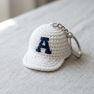 Può includere: Un portachiavi a forma di cappellino da baseball lavorato all'uncinetto in bianco con una lettera A ricamata in blu scuro sul davanti. Il cappellino in miniatura è realizzato in filato ed è fissato a un anello in metallo argentato.