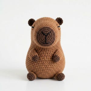 Peut inclure: Un jouet en peluche capybara crocheté marron avec un corps arrondi, de petites oreilles marron foncé et des pattes marron foncé. Le visage présente un museau circulaire marron plus foncé avec des détails noirs brodés pour le nez et la bouche, et des yeux fermés.