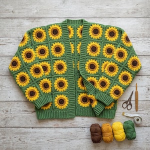 Könnte beinhalten: Handgestrickte grüne Strickjacke mit Sonnenblumenmuster. Die Strickjacke hat einen klassischen offenen Schnitt mit geripptem Saum und Bündchen. Die Sonnenblumen sind gelb und braun. Häkelnadel, Schere und Garn liegen daneben.