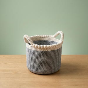 May include: A handmade crochet basket in gray and cream. The basket has a cylindrical shape with two arched handles. The rim is cream-colored with a decorative beaded edge. The interior is a lighter gray.