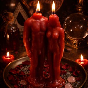 May include: Two red wax candles shaped like human figures standing side by side on a round metal tray. Each candle has a lit wick at the top. The tray is decorated with scattered dried rose petals, small grey seeds, and smooth light pink stones.