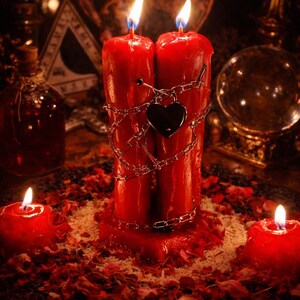 May include: Two tall red pillar candles joined together, wrapped in a silver-toned metal chain with a heart-shaped padlock attached. The candles are lit, with two smaller red votive candles placed on either side amidst scattered red flower petals.