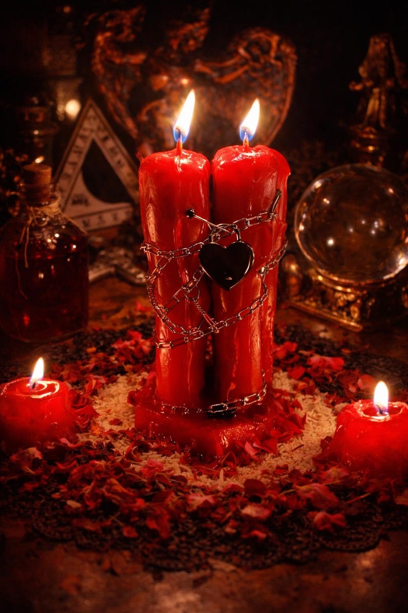 May include: Two tall red pillar candles joined together, wrapped in a silver-toned metal chain with a heart-shaped padlock attached. The candles are lit, with two smaller red votive candles placed on either side amidst scattered red flower petals.