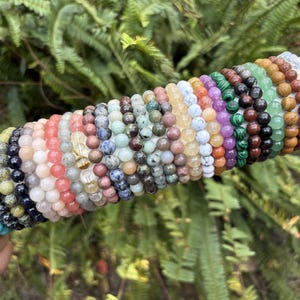May include: A collection of beaded bracelets in various colors, including turquoise, black, pink, and green. The bracelets are made of natural stones and are displayed on a white cylindrical object against a backdrop of green foliage.