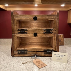 May include: A two-tier rustic bread box made of stained wood. The bread box has two compartments with black hardware and a black handle. A tag with the words "HANDMADE OAK" is attached to the bread box.