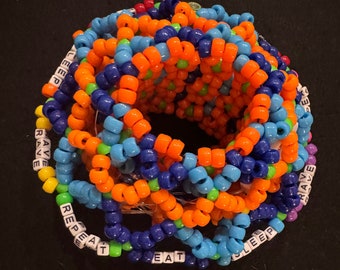 Peut inclure: Un grand bracelet kandi sphérique dans une variété de couleurs, dont l'orange, le bleu et le vert. Le bracelet comporte les mots "EAT", "SLEEP", "RAVE" et "REPEAT" épelés en perles de lettres blanches. Les perles sont enfilées pour former un accessoire portable.