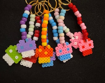 Puede incluir: Una colección de llaveros de cuentas coloridas. Cada llavero presenta un anillo dorado y una hebra de cuentas de varios colores, que conduce a un dije pixelado. Los dijes tienen forma de cupcakes y otros diseños, en tonos verdes, naranjas, azules y rosas.