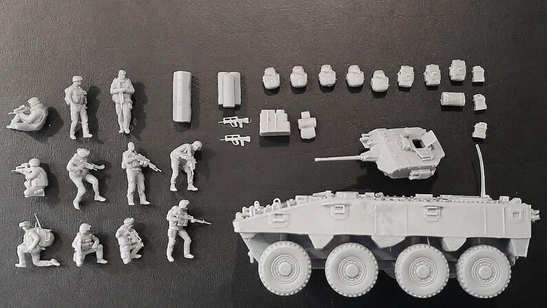Op de afbeelding: Een verzameling ongeverfde grijze hars modelbouwset onderdelen, waaronder negen soldaatfiguren in diverse tactische houdingen, een pantservoertuigchassis met meerdere wielen, een losse koepel en een assortiment aan kleine uitrusting en wapens.