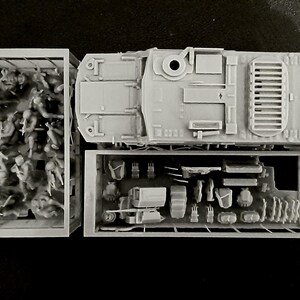 May include: A gray plastic model kit of a military vehicle, with separate compartments for soldiers and equipment. The top view shows the vehicle's details, including hatches and vents. The kit includes miniature soldiers and various accessories.