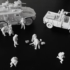 Op de afbeelding: Een verzameling ongeverfde lichtgrijze hars miniatuurfiguren, waaronder een militair pantservoertuig, een vrachtwagen en verschillende soldaten in diverse tactische houdingen, sommige staand en andere bezig met uitrusting op de grond.