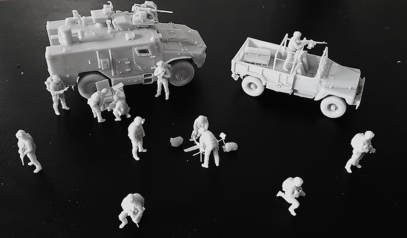 Op de afbeelding: Een verzameling ongeverfde lichtgrijze hars miniatuurfiguren, waaronder een militair pantservoertuig, een vrachtwagen en verschillende soldaten in diverse tactische houdingen, sommige staand en andere bezig met uitrusting op de grond.