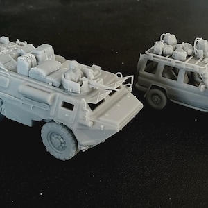 Op de afbeelding: Twee ongeverfde grijze hars schaalmodellen van militaire voertuigen. De ene is een gepantserd troepentransportvoertuig met een gemonteerd machinegeweer en vracht, de andere is een terreinwagen met een dakdrager vol met diverse uitrusting.