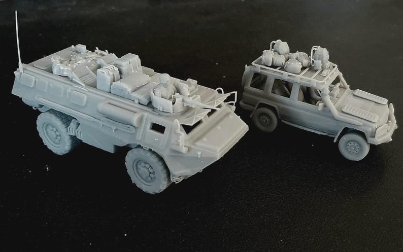 Op de afbeelding: Twee ongeverfde grijze hars schaalmodellen van militaire voertuigen. De ene is een gepantserd troepentransportvoertuig met een gemonteerd machinegeweer en vracht, de andere is een terreinwagen met een dakdrager vol met diverse uitrusting.