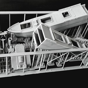 Peut inclure: Un kit de mod&egrave;le imprim&eacute; en 3D gris clair contenu dans un cadre de support rectangulaire. Le kit comprend une coque de bateau, une section de superstructure rectangulaire et divers petits composants d&eacute;taill&eacute;s fix&eacute;s &agrave; des supports verticaux.