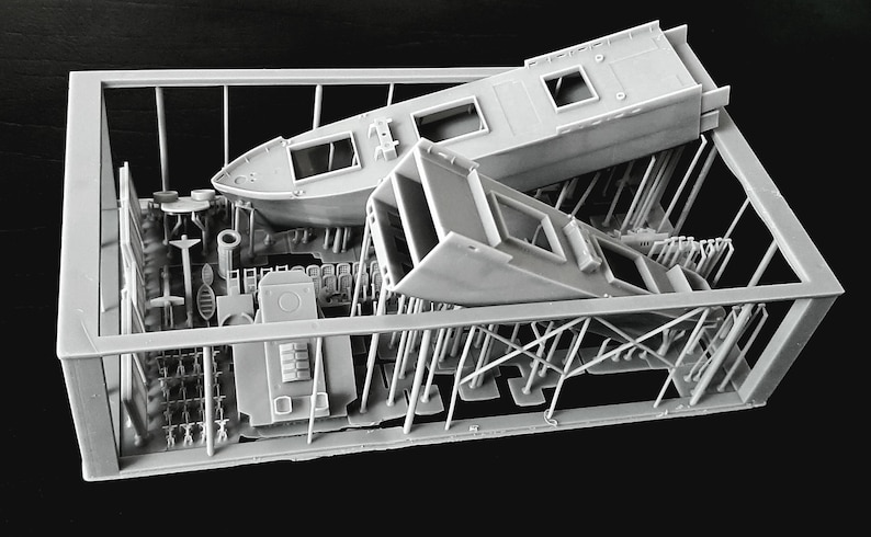 Peut inclure: Un kit de mod&egrave;le imprim&eacute; en 3D gris clair contenu dans un cadre de support rectangulaire. Le kit comprend une coque de bateau, une section de superstructure rectangulaire et divers petits composants d&eacute;taill&eacute;s fix&eacute;s &agrave; des supports verticaux.
