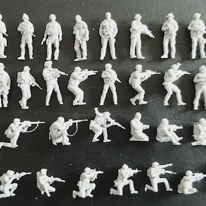 Op de afbeelding: Een verzameling onbeschilderde witte plastic miniatuursoldaatjes in verschillende gevechtshoudingen. De set bevat staande, knielende en hurkende figuren, allemaal uitgerust met geweren of andere uitrusting, gerangschikt in vier horizontale rijen.