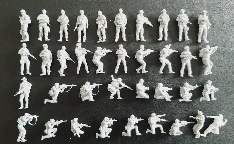 Op de afbeelding: Een verzameling onbeschilderde witte plastic miniatuursoldaatjes in verschillende gevechtshoudingen. De set bevat staande, knielende en hurkende figuren, allemaal uitgerust met geweren of andere uitrusting, gerangschikt in vier horizontale rijen.