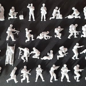 Op de afbeelding: Een verzameling kleine, witte plastic militaire beeldjes in verschillende actieposities. De figuren beelden soldaten uit in verschillende houdingen, waaronder knielen, wapens richten en uitrusting dragen. Op een donkere ondergrond.