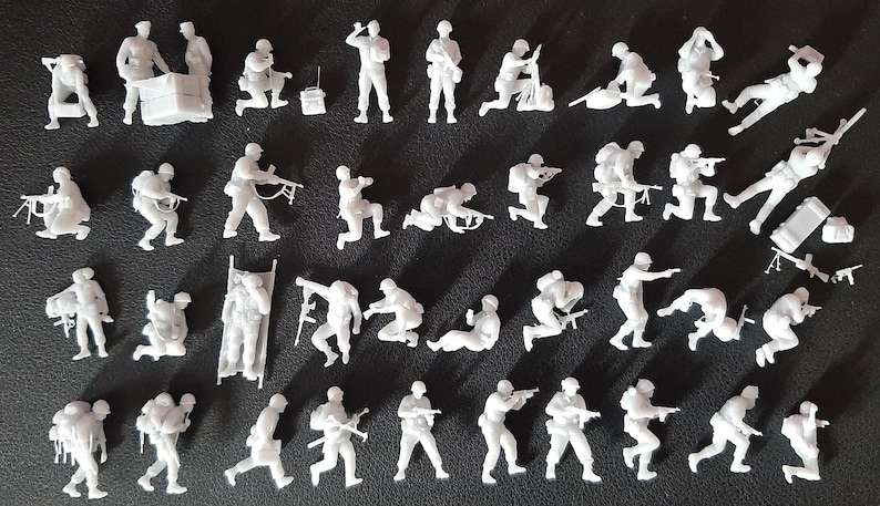 Op de afbeelding: Een verzameling kleine, witte plastic militaire beeldjes in verschillende actieposities. De figuren beelden soldaten uit in verschillende houdingen, waaronder knielen, wapens richten en uitrusting dragen. Op een donkere ondergrond.