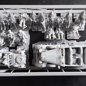 Pu&ograve; includere: Un kit di modelli in plastica grigia contenente figure in miniatura di soldati e due veicoli. Le figure sono in varie pose e i veicoli sembrano essere modelli di trasporto militare. Il kit &egrave; in una cornice rettangolare.