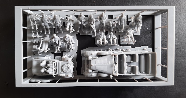 Pu&ograve; includere: Un kit di modelli in plastica grigia contenente figure in miniatura di soldati e due veicoli. Le figure sono in varie pose e i veicoli sembrano essere modelli di trasporto militare. Il kit &egrave; in una cornice rettangolare.