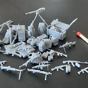 Puede incluir: Colecci&oacute;n de modelos militares grises en miniatura, que incluyen rifles, ametralladoras, mochilas, cajas y un mortero con tr&iacute;pode. Se incluye una cerilla para la escala, y el texto "x2" es visible.