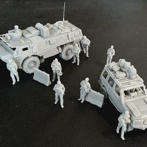 Op de afbeelding: Een set onbeschilderde grijze hars miniatuurmodellen met een militair gepantserd troepentransportvoertuig en een terreinwagen, beide beladen met uitrusting, vergezeld door diverse staande soldaatfiguren met geweren en verplaatsbare barri&egrave;res.