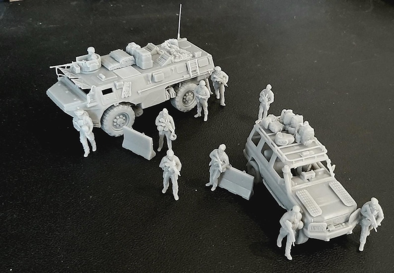 Op de afbeelding: Een set onbeschilderde grijze hars miniatuurmodellen met een militair gepantserd troepentransportvoertuig en een terreinwagen, beide beladen met uitrusting, vergezeld door diverse staande soldaatfiguren met geweren en verplaatsbare barri&egrave;res.
