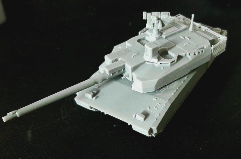 Op de afbeelding: Een lichtgrijs, onbeschilderd schaalmodel van een gevechtstank met een lange loop, een draaibare koepel met een gemonteerd machinegeweer en gedetailleerde rompbeplating. Het model is vervaardigd uit een eenkleurig harsmateriaal.