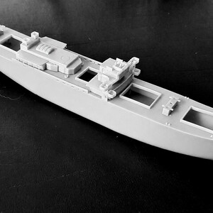 Peut inclure: Une maquette de navire en plastique gris clair pr&eacute;sentant un pont d&eacute;taill&eacute; avec de multiples ouvertures rectangulaires, une superstructure centrale et des &eacute;l&eacute;ments structurels en relief le long de la coque. Le mod&egrave;le non peint affiche un design naval &eacute;lanc&eacute;.