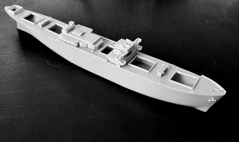 Peut inclure: Une maquette de navire en plastique gris clair pr&eacute;sentant un pont d&eacute;taill&eacute; avec de multiples ouvertures rectangulaires, une superstructure centrale et des &eacute;l&eacute;ments structurels en relief le long de la coque. Le mod&egrave;le non peint affiche un design naval &eacute;lanc&eacute;.