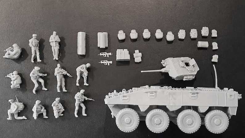 Op de afbeelding: Een verzameling onbeschilderde miniatuur militaire modellen, waaronder soldaten in verschillende poses, een tank en extra uitrusting. De modellen zijn gemaakt van een lichtgrijs materiaal en zijn gerangschikt op een donkere ondergrond. De tank heeft acht wielen.