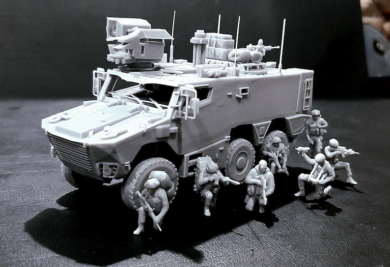 May include: A gray model of a military armored vehicle with multiple antennas and a mounted weapon system. Miniature soldiers are positioned around the vehicle, some holding rifles. The model is detailed and appears to be a scale replica.