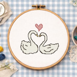 May include: Cross-stitch embroidery in a wooden hoop featuring two white swans forming a heart shape with a pink heart above. The design is on white fabric, set against a blue and white checkered background. Embroidery supplies are visible.