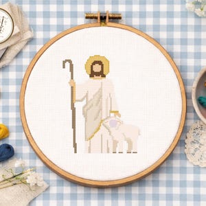 May include: A cross-stitch embroidery hoop featuring a religious figure with a staff, a halo, and a lamb. The design is in shades of white, beige, and brown, set against a white fabric. The wooden hoop is visible, and the background is a blue and white checkered pattern.